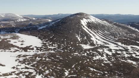 Aerial-drone-flying-out-over-a-cliff-face-towards-a-dramatic-mountain-and-moorland-landscape-covered-with-patches-of-snow-and-clear-blue-skies-near-Ben-Macdui-in-the-Cairngorms-National-Park,-Scotland