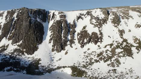 Aerial-drone-footage-flying-slowly-above-a-beautiful-frozen-Loch-Etchachan-towards-a-dramatic-and-imposing-ice-covered-rock-face-and-cliff-with-steep-snow-filled-gullies