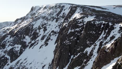 Aerial-drone-footage-rising-and-tilting-to-reveal-a-mountain-cliff-face-and-snow-filled-gullies-near-Ben-Macdui-in-the-Cairngorms-National-Park,-Scotland-as-sunshine-reflects-off-of-snow-on-a-ridge