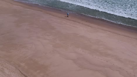 Slow-Drone-Fly-Over-Male-Fisherman-Walking-Alone-Beach-Shoreline-Waves-Crashing