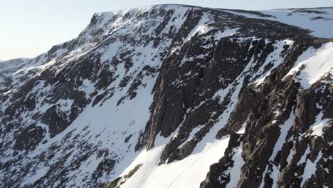 Aerial-drone-footage-flying-towards-a-steep-mountain-cliff-face-and-snow-filled-gullies-near-Ben-Macdui-in-the-Cairngorms-National-Park,-Scotland-as-sunshine-reflects-off-of-a-snow-covered-ridge