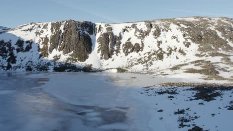 Aerial-drone-footage-flying-close-above-a-beautiful-frozen-Loch-Etchachan-towards-a-dramatic-and-imposing-ice-covered-rock-face-and-cliff-with-steep-snow-filled-gullies