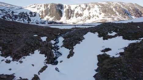 Aerial-drone-footage-following-an-icy-river-towards-a-beautiful-frozen-Loch-Etchachan-and-imposing-ice-covered-rock-face-and-cliff-with-steep-snow-filled-gullies