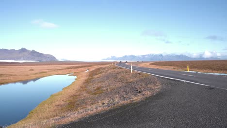 Coches-Circulando-Por-Una-Carretera-Asfaltada-Junto-A-Un-Pequeño-Lago-En-Islandia