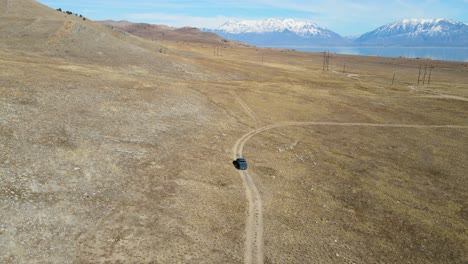 Camión-Conduciendo-Por-Un-Camino-De-Tierra-En-Un-Pasto-Seco-Que-Revela-Un-Lago-Y-Montañas-Cubiertas-De-Nieve-En-La-Distancia