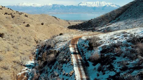 A-dirt-road-along-a-canyon-leading-out-to-a-valley-with-a-lake-and-snow-capped-mountains---aerial-flyover