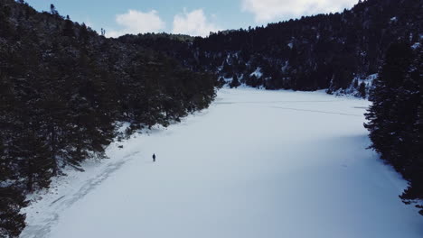 Aerial-shot-following-a-man-walking-on-frozen-lake-covered-of-snow,-at-the-background-a-mountain-full-of-snow,-revealing-vast-high-altitude-forest