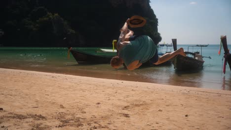 Strong-muscled-athletic-european-man-does-a-side-somersault-on-the-beautiful-sandy-beach-of-Krabi-while-touristic-Thai-boats-are-moored-in-the-background