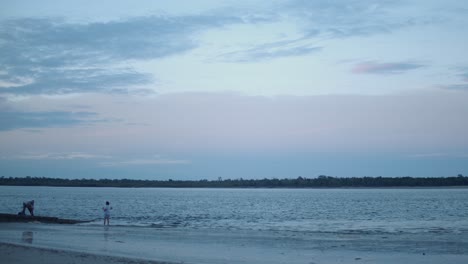 Two-elderly-people-stand-on-the-shore-of-a-reflective-beach-fishing-under-the-fluffy,-white-clouds-floating-overhead-being-bathed-in-pastel-blue-and-pink-colours-from-the-setting-sun-at-blue-hour