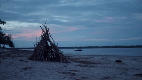 A-teepee-made-of-various-sized-sticks-sits-on-the-peaceful-beach-as-the-retreating-sun-spills-pastel-colours-of-pink-and-blue-into-the-clouds,-ocean-and-water-whilst-a-boat-floats-along-the-shoreline