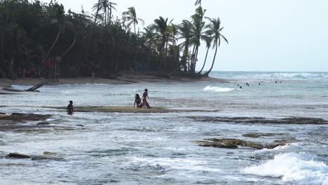 Mother-and-two-children-sit-at-tropical-beach-over-the-rocky-shore,-Handheld-wide-shot