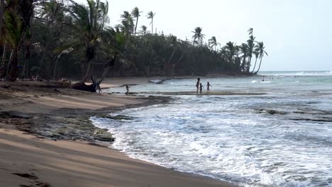 Mother-and-three-children-play-at-tropical-beach-over-the-rocky-shore,-Handheld-wide-shot