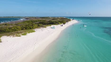 Man-Kitesurf-Jump-on-turquoise-caribbean-sea,-drone-shot-along-Kite-camp-on-white-sand-beach