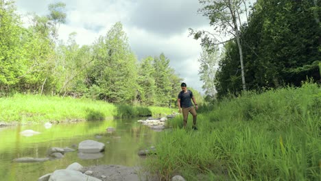 Lone-modern-male-hiker-in-the-wilderness-searching-overgrown-riverbank-in-the-middle-of-a-forest-landscape-with-rocks-and-lush-green-summer-foliage