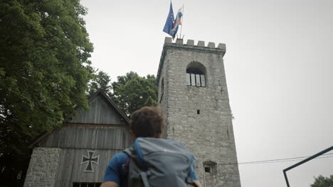 Camera-tracking-from-behind-a-young-hiker-walkig-towards-the-tower-where-two-flags-flutter-in-the-wind