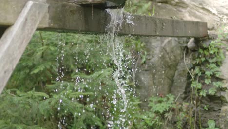 Wasser,-Das-Von-Einem-Holzbrett-Aus-Einer-Historischen-Mühle-Mit-Einem-Verschwommenen-Wald-Im-Hintergrund-Herunterläuft