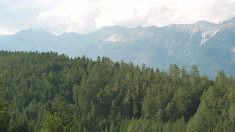 Paisaje-Escénico-De-Bosques-De-Coníferas-En-Una-Colina-Con-Una-Cordillera-Alpina-En-El-Fondo-Con-Nubes-Texturizadas-Alrededor-En-Un-Soleado-Día-De-Otoño
