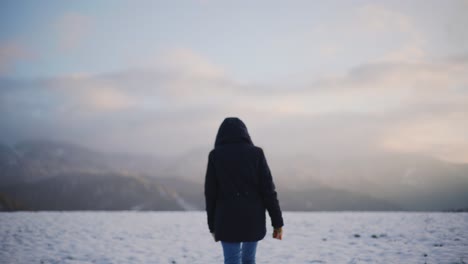 Beautiful-young-woman-walking-through-a-snow-covered-field-in-winter,-wearing-a-blue-hooded-jacket