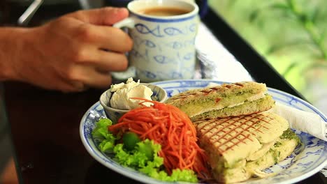 Selektiver-Fokus-Auf-Einen-Teller-Mit-Veganen-Panini-Zum-Frühstück,-Während-Eine-Person-Im-Hintergrund-Kaffee-Trinkt