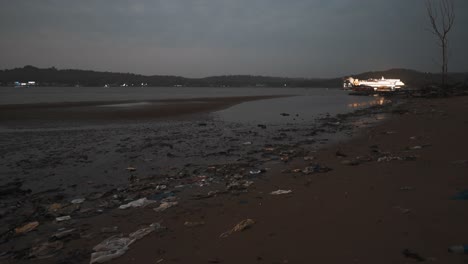 An-evening-shot-overlooking-the-Mandovi-River-during-low-tide,-the-sand-on-the-riverbank-covered-in-plastic-waste-and-trash-which-is-harmful-to-the-natural-environment,-Goa,-India