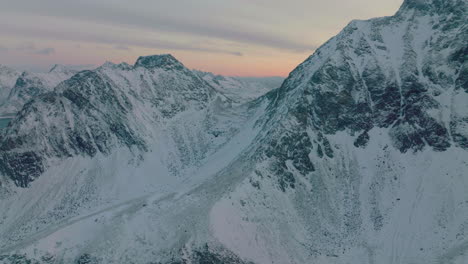 Stunning-Lofoten-frozen-wintry-mountain-range-slopes-aerial-view-flying-towards-glacial-summit-at-sunrise