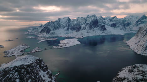 Flying-over-extreme-snowy-Lofoten-Reine-mountain-peaks-overlooking-picturesque-wintry-blue-ocean
