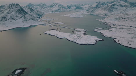 Nordic-Ramberg-village-on-glacial-lake-fjord-aerial-view-above-snowy-mountain-range