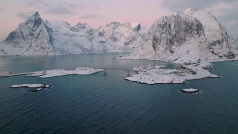 Descending-aerial-view-of-snowy-Moskenes-mountains-to-scenic-Reine-fishing-village-at-sunrise