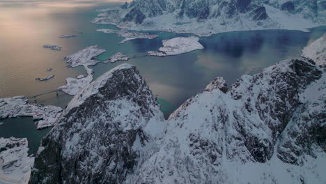 Fliegende-Lofoten-Schneebedeckte-Reine-Berggipfel-Mit-Blick-Auf-Den-Eisblauen-Ozean-Sonnenuntergang-Himmel