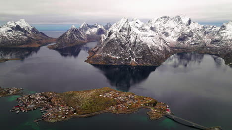 Aerial-view-orbiting-glacial-Moskenes-mountains-and-Reine-fishing-village-reflecting-in-winter-ocean-at-sunrise