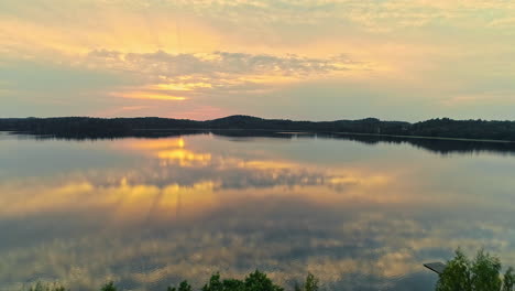 Luftaufnahme-Des-Ruhigen-Reflektierenden-Sees-Mit-Orangefarbenen-Sonnenuntergangsstrahlen-Und-Wolken