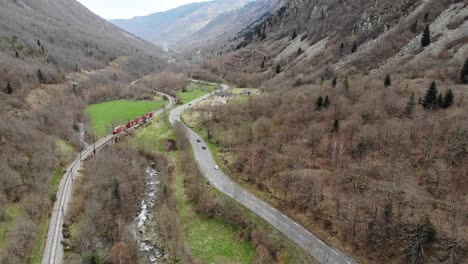 Aerial:-passenger-train-in-a-valley-by-a-river-and-a-road-with-light-traffic-in-the-Pyrenees,-southern-France