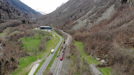 Aerial:-passenger-train-in-a-valley-by-a-road-with-light-traffic-in-the-Pyrenees,-southern-France