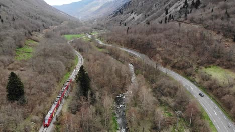 Aerial:-passenger-train-in-a-valley-by-a-river-and-a-road-with-light-traffic-in-the-Pyrenees,-southern-France