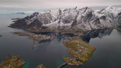 Moskenesøya-snowy-mountain-reflections-in-ocean-aerial-view-flying-over-Reine-Norway-fishing-village-at-sunrise