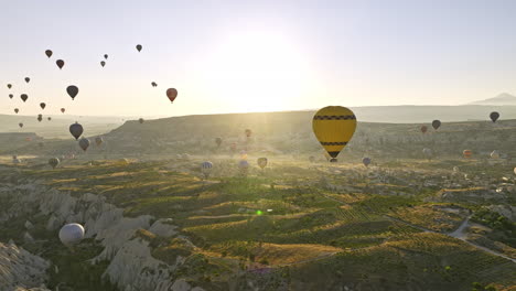 Göreme-Turkey-Aerial-v65-sunrise-landscape-view-capturing-open-terrain-with-extraordinary-rock-formations,-colorful-hot-air-balloons-and-glowing-sun-in-the-sky---Shot-with-Mavic-3-Cine---July-2022