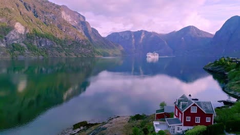 Tiny-little-lakeside-house-with-a-view-of-majestic-mountain-landscape,-river-Flåmsdalen-and-cruise-at-far,-Norway