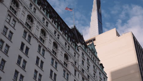 Low-Angle-View-of-Plaza-Hotel-Building-Facade-in-New-York-City-Manhattan-Usa,-American-Flag-on-Top-of-Building,-Urban-Architecture-and-Modern-Downtown-Tower-in-Background
