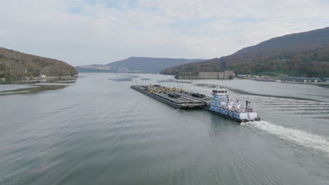 Aerial-footage-of-a-boat-pushing-a-large-industrial-barge-through-the-Tennessee-River-past-Historic-Hales-Dam-Powerhouse-in-Autumn