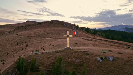 Luftaufnahme-Nach-Hinten-Von-Beleuchtungskreuz-Mit-Schwenkender-Rumänischer-Flagge-Auf-Dem-Gipfel-Des-Mount-Dichiu-Bei-Sonnenuntergang