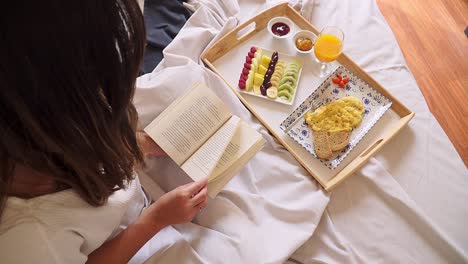 Young-woman-reading-a-book-on-a-hotel-room-bed-while-having-healthy-breakfast