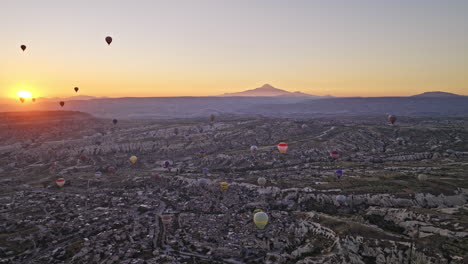 Göreme-Turkey-Aerial-v50-panoramic-panning-view,-flyover-old-village-town-capturing-beautiful-hot-air-balloons-flying-in-the-sky-with-sun-rising-above-the-horizon---Shot-with-Mavic-3-Cine---July-2022