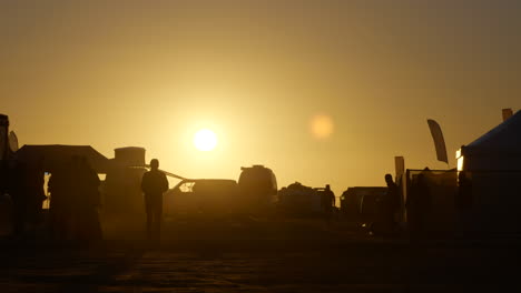 Silhouetten-Von-Menschen-Und-Fahrzeugen-Im-Staubigen-Dakar-Rallye-Camp-Unter-Glühendem-Sonnenuntergang