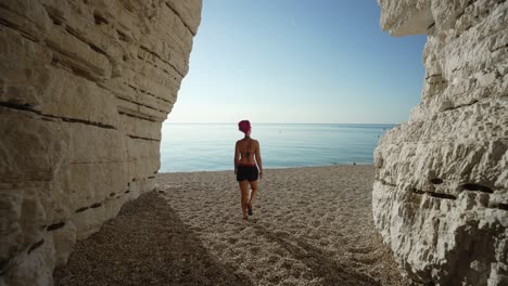 Woman-followed-from-behind-walking-out-of-a-cave-on-to-the-beach-towards-the-blue-sea-in-Italy