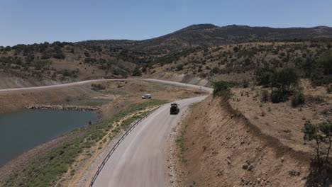 Drone-following-red-trucktor-slowly-driving-a-road-in-turkey-karaman-between-blue-lake-and-mountains-at-a-sunny-summer-though-curves