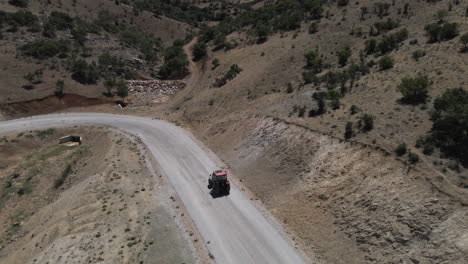 Drohne-Hinter-Rotem-Trucktor,-Der-Langsam-Eine-Straße-In-Der-Türkei-Karaman-Zwischen-Feldern-Und-Bergen-An-Einem-Sonnigen-Sommer-Durch-Kurven-Fährt
