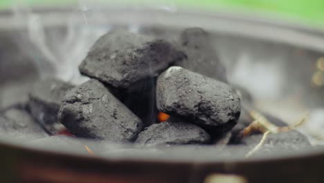 Charcoal-laying-in-a-black-pan-smoking,-starting-to-glow-on-a-summer-day-with-blurred-grass-in-the-background