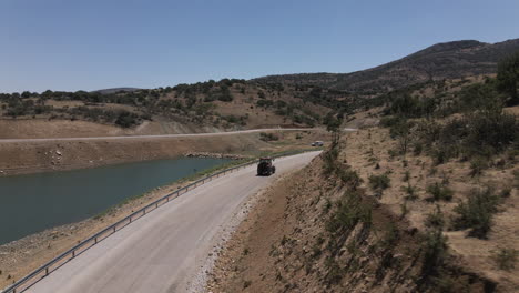 Drone-following-red-trucktor-slowly-driving-a-road-in-turkey-karaman-between-blue-lake-and-mountains-at-a-sunny-summer-though-curves