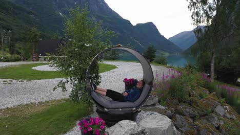 Cute-girl-smiling-while-relaxing-inside-circular-pipe-bed-in-spectacular-landscape-at-Yrineset-Olden-glacial-lake---Aerial-flying-through-pipe-before-ascending-to-reveal-majestic-mountain-landscape