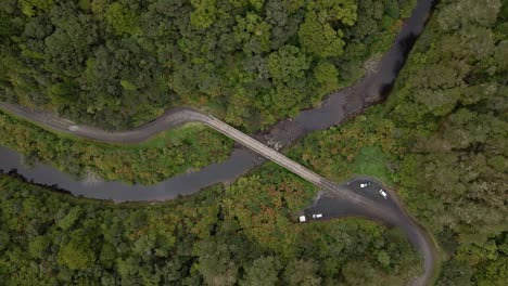 Descending-top-view-of-a-one-lane-bridge-crossing-a-brown-river-in-a-dense-rainforest